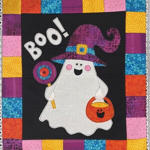 May include: A white ghost wearing a purple witch hat with a yellow buckle and holding a lollipop and a trick-or-treat bag. The background is a black fabric with a colourful patchwork border. The text "BOO!" is written in white on the black fabric.