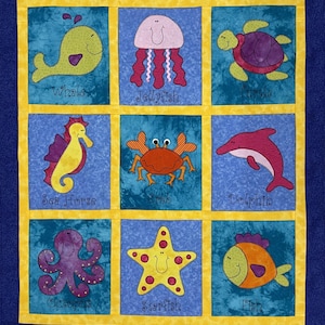 May include: A colourful quilt with nine squares featuring different sea creatures. Each square has a different coloured background and a different sea creature in the centre. The sea creatures include a whale, jellyfish, turtle, seahorse, crab, dolphin, octopus, starfish, and fish. Each square also has the name of the sea creature written below it.