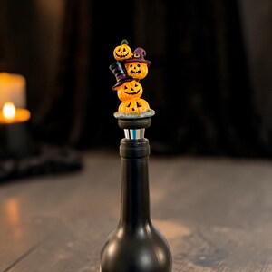 May include: A bottle stopper featuring a stack of four orange jack-o'-lanterns. The top pumpkin is small, the second wears a purple witch hat, the third wears a black top hat, and the base pumpkin sits on a gray stone-like platform.