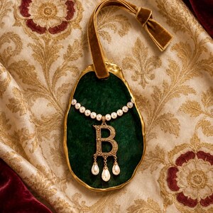 May include: A gold-rimmed, dark green oval ornament featuring a gold-toned letter B pendant adorned with three dangling white pearls. A string of small white pearls encircles the top of the letter, and a brown velvet ribbon is attached to the top.