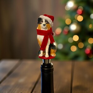 May include: A decorative wine bottle stopper featuring a dog figurine wearing a Santa hat and a red scarf. The dog is sitting on a red base, with a black bottle neck below. The background is blurred with a Christmas tree.
