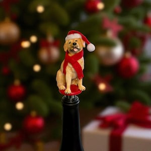 May include: A festive wine bottle stopper featuring a golden Labrador Retriever wearing a Santa hat and a red scarf. The dog figurine sits atop a red base, perfect for holiday decor.