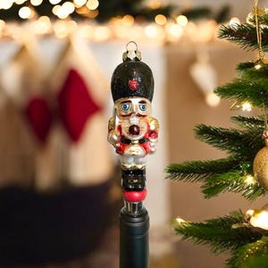 May include: A decorative bottle stopper featuring a nutcracker soldier design. The ornament has a black hat with a red accent, a red and gold uniform, and is attached to a black bottle. The background includes a Christmas tree and festive lights.