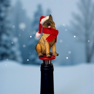 May include: A German Shepherd wine bottle stopper. The dog figurine wears a red Santa hat and scarf. The dog is tan and black, sitting on a red base. The bottle stopper is on top of a black wine bottle.