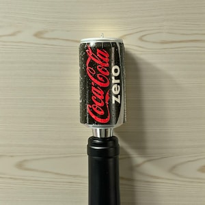 May include: A Coca-Cola Zero can bottle stopper. The can is black with red Coca-Cola branding and white "Zero" text. The stopper has a silver base and a black bottle neck.