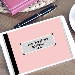 May include: A pink digital tablet with a white border and a black stripe on the left side. The screen displays a gray rectangle with the text "Grace Through Faith Life Planner 2020".