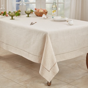 May include: A rectangular beige linen tablecloth with a hemstitched border detail. The table is set with a glass bottle, a wooden bowl containing fruit, two small green glass bowls, a white plate, a fork, a knife, and two small bird figurines.