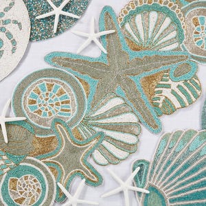 May include: A collection of circular and shaped beaded placemats and table runners featuring coastal motifs including crabs, starfish, and various seashells in shades of turquoise, gold, white, and tan, accented by small white starfish figurines.
