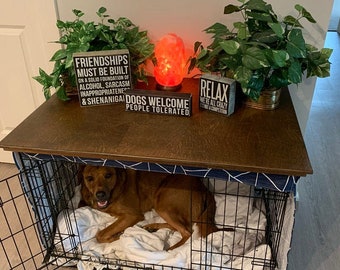 May include: A brown dog is lying in a black metal dog crate with a white blanket. The crate is on a wooden table with a blue and white patterned fabric cover. There are three wooden signs on the table that say "Friendships must be built on a solid foundation of alcohol, sarcasm, inappropriateness & shenanigans", "Dogs welcome people tolerated", and "Relax we're all crazy". There is a small orange Himalayan salt lamp on the table.
