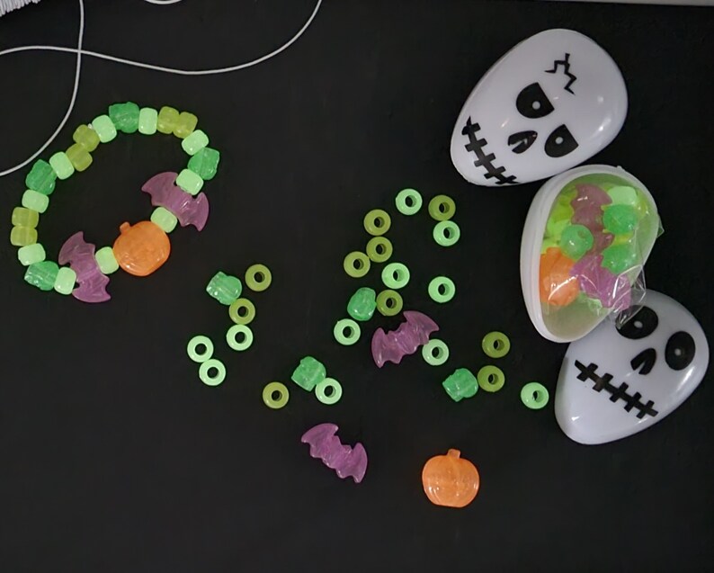 May include: A set of glow-in-the-dark Halloween-themed beads, including bats, pumpkins, and ghosts. The beads are in a variety of colors, including green, orange, and purple. The beads are in a plastic container shaped like a skull.