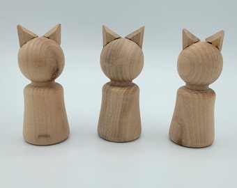 May include: Three wooden cat figurines. Each figurine has a round head with pointed ears and a cylindrical body. The wood has a natural, light brown color. The figurines are arranged in a row against a white background.
