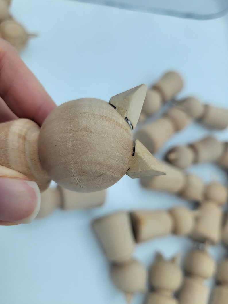 May include: A close-up of a wooden doll with a round head and two pointed ears. The doll is made of unfinished wood and is ready to be painted or decorated.