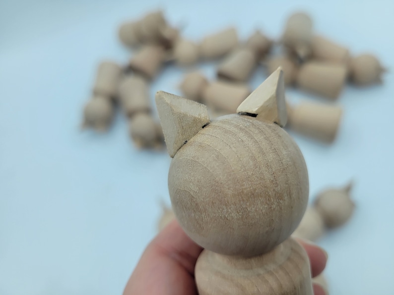May include: A wooden toy cat with triangular ears. The cat is light brown and has a smooth, rounded body.