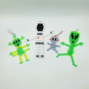 Puede incluir: Cuatro colgantes espaciales de cuentas. Incluye dos extraterrestres verdes, un robot gris y un astronauta blanco. Cada colgante está hecho de pequeñas cuentas de colores y tiene un cierre plateado para sujetar.