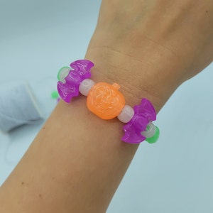 May include: A purple and green Halloween themed bracelet with a pumpkin charm. The bracelet is made of plastic beads and is worn on a person's wrist.