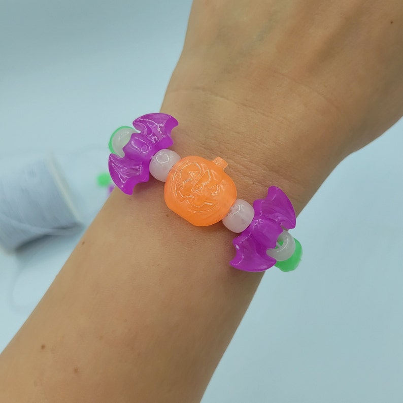 May include: A purple and green Halloween themed bracelet with a pumpkin charm. The bracelet is made of plastic beads and is worn on a person's wrist.