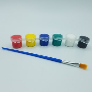 May include: A set of six small paint pots in red, yellow, blue, green, white, and black, each with a clear lid. A blue-handled paintbrush with a rectangular, tan-colored brush head lies below the paint pots.