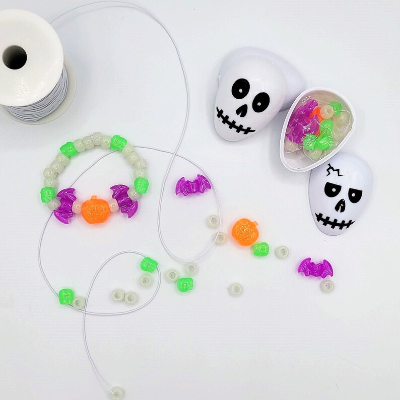 May include: A white string with glow-in-the-dark Halloween beads, including pumpkins, bats, and skulls. The beads are strung on a white string and are arranged in a circle. There are also some loose beads scattered around the string.