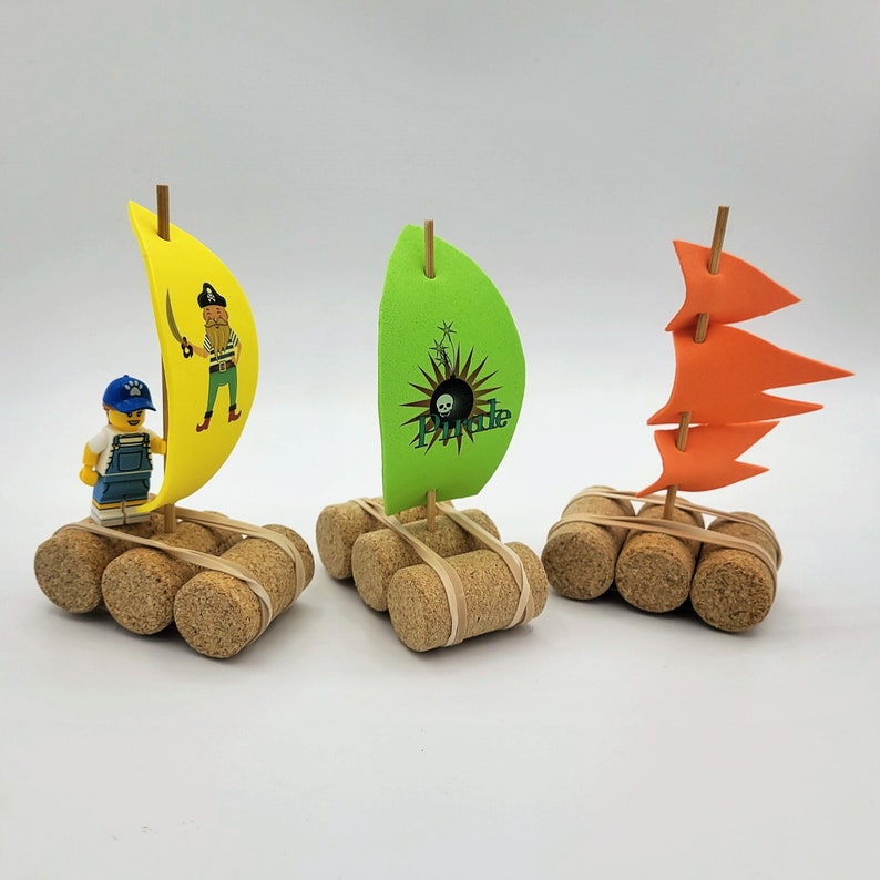 May include: Three miniature rafts made of cork with colorful paper sails. The rafts are decorated with a pirate, a skull and crossbones, and a red sail.