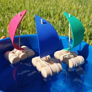 May include: Three toy sailboats made from cork and foam are floating in a blue plastic tub of water. The sailboats are red, blue, and green.