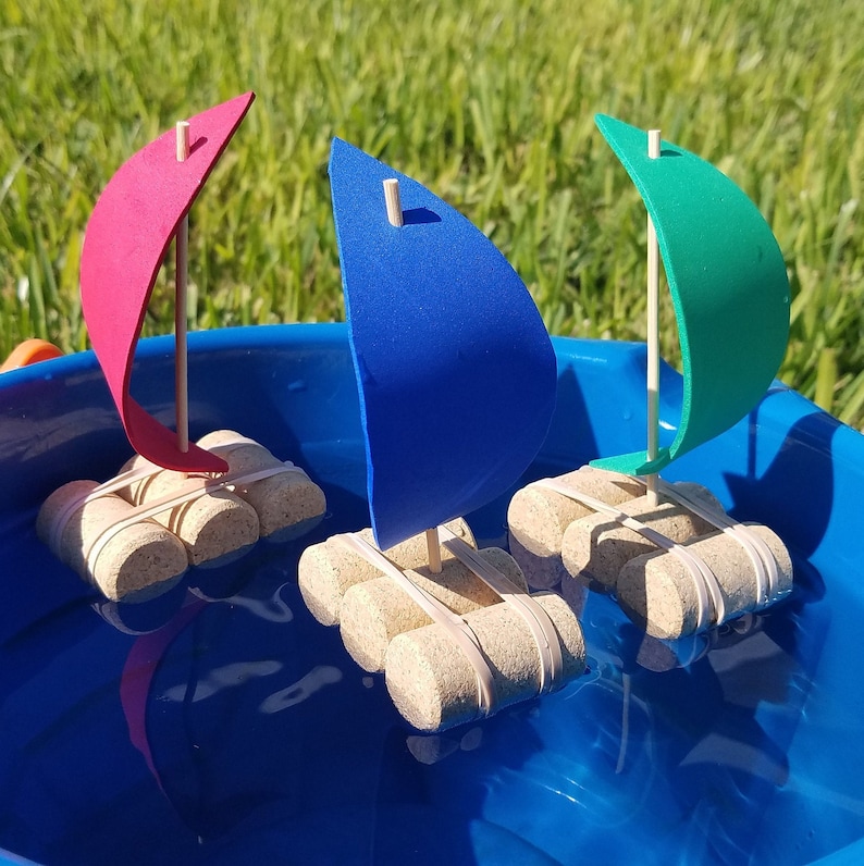May include: Three toy sailboats made from cork and foam are floating in a blue plastic tub of water. The sailboats are red, blue, and green.