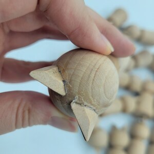 May include: Unfinished wooden doll head with a pointed, triangular shape at the top. The doll head is made of light brown wood and is being held by a hand.