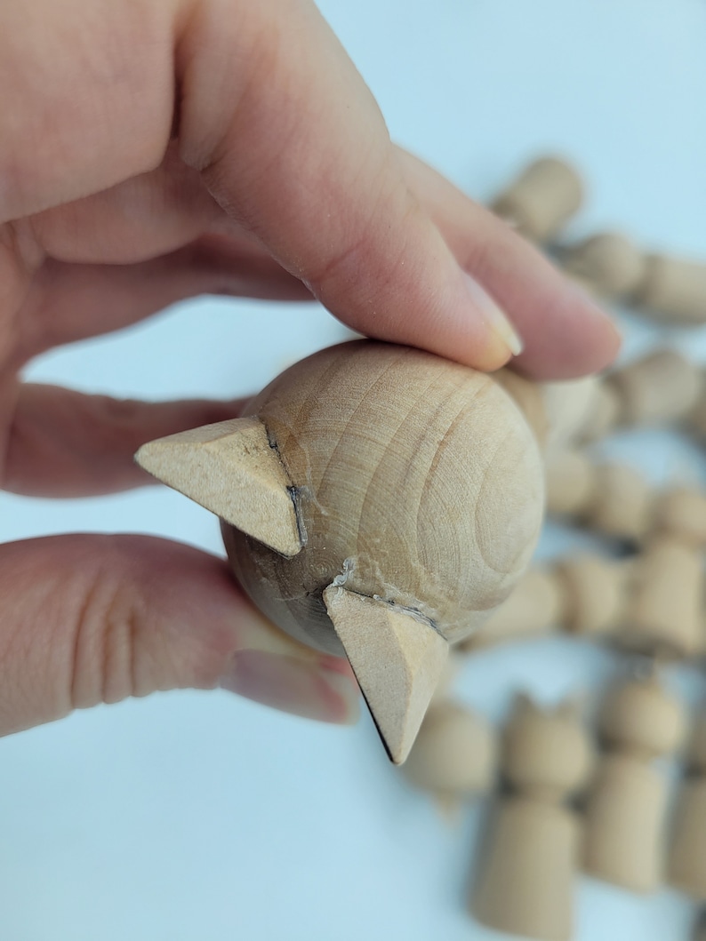 May include: Unfinished wooden doll head with a pointed, triangular shape at the top. The doll head is made of light brown wood and is being held by a hand.