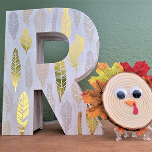 Peut inclure: Une lettre en bois R avec un motif de plumes dor&eacute;es et blanches, &agrave; c&ocirc;t&eacute; d'une dinde artisanale faite d'une tranche de bois avec des yeux globuleux et des feuilles d'automne.