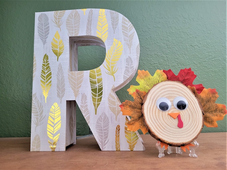 Peut inclure: Une lettre en bois R avec un motif de plumes dor&eacute;es et blanches, &agrave; c&ocirc;t&eacute; d'une dinde artisanale faite d'une tranche de bois avec des yeux globuleux et des feuilles d'automne.