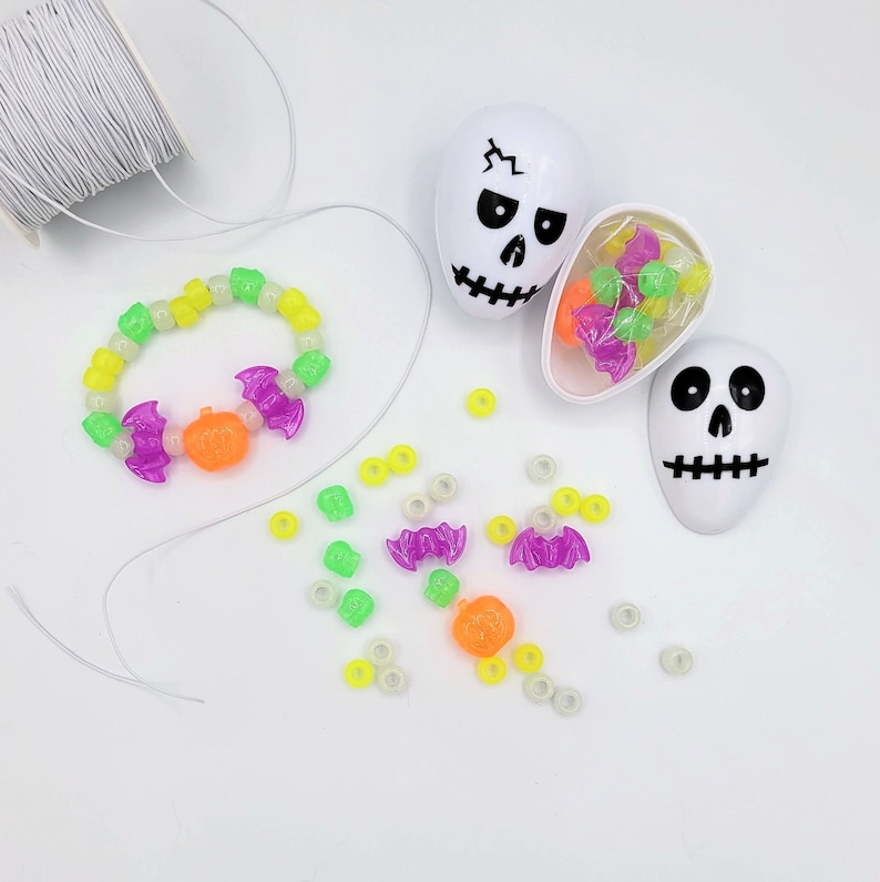 May include: A white plastic skull-shaped container with a black face and a purple and green glow-in-the-dark beaded bracelet. The bracelet has a pumpkin and bat charm. There are also loose glow-in-the-dark beads in the image.