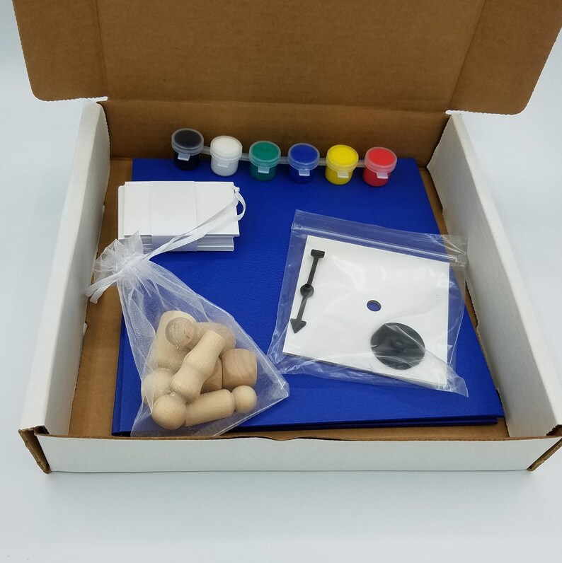 Puede incluir: Una estera de artesan&iacute;a azul con un juego de seis botes de pintura, una tarjeta blanca, una bolsa de pl&aacute;stico transparente con una flecha negra y un c&iacute;rculo negro, y una pila de clavijas de madera.