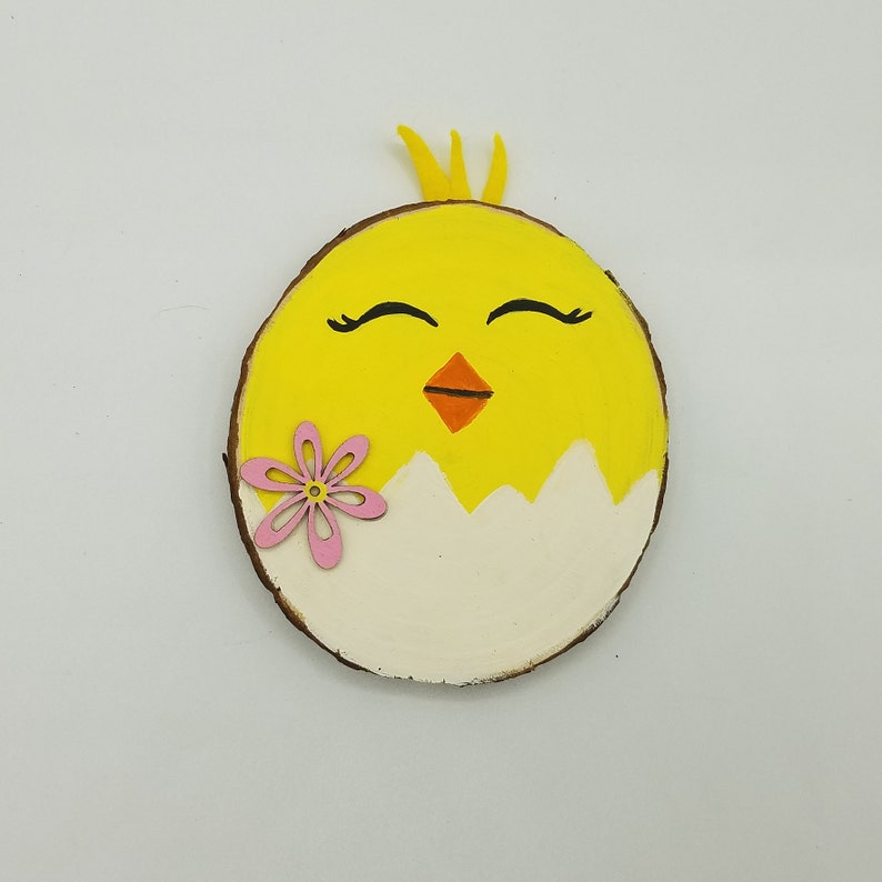 Op de afbeelding: Een gele kuiken geschilderd op een ronde houten schijf met een roze bloem. De kuiken heeft gesloten ogen en een kleine oranje snavel.
