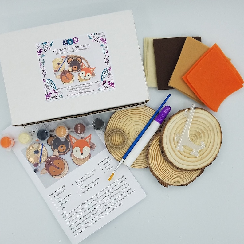 wood slices, glue, paint, paint, paint brush, pieces of felt, and an instruction manual are placed around a white box with a photo of a bear, deer, and fox ornament