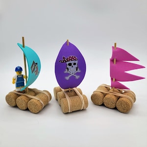 May include: Three miniature pirate ships made from wine corks and foam. The ships are decorated with colorful sails and pirate-themed designs. One ship has a blue sail with the word "Ahoy" on it, another has a purple sail with a skull and crossbones, and the third has a pink sail.
