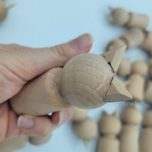 May include: A close-up of a wooden toy cat with a light brown finish. The cat is unfinished and has a simple design. The cat is being held in a hand.