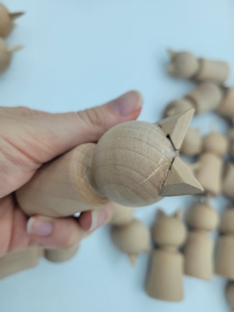 May include: A close-up of a wooden toy cat with a light brown finish. The cat is unfinished and has a simple design. The cat is being held in a hand.