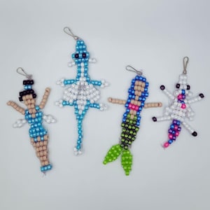 May include: Four beaded charms in the shape of a mermaid, lizard, ballerina, and unicorn. The charms are made of small, colorful beads strung together with silver-colored clasps. The mermaid has a green tail, the lizard is blue and white, the ballerina is blue and tan, and the unicorn is white and pink.