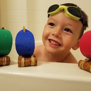 May include: A young child wearing yellow goggles is in a bathtub with three homemade toy boats. The boats are made from cork and foam and have green, blue, and red sails.