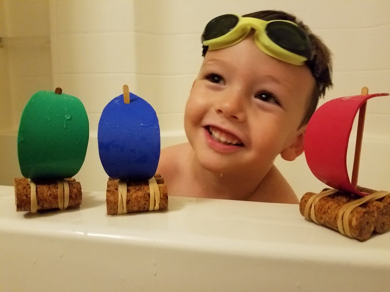 May include: A young child wearing yellow goggles is in a bathtub with three homemade toy boats. The boats are made from cork and foam and have green, blue, and red sails.