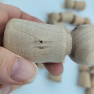 May include: Close-up of a wooden doll head with two small holes drilled into the top. The doll head is light brown and has a smooth, unfinished surface.