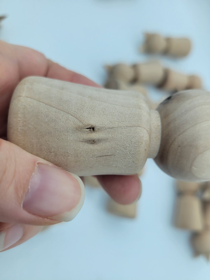May include: Close-up of a wooden doll head with two small holes drilled into the top. The doll head is light brown and has a smooth, unfinished surface.