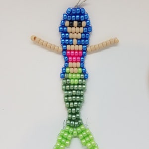 Peut inclure: Un charme de sir&egrave;ne en perles avec un haut bleu, un bikini rose et une queue verte. Le charme est fait de petites perles en plastique et a une boucle en haut pour l'accrocher.