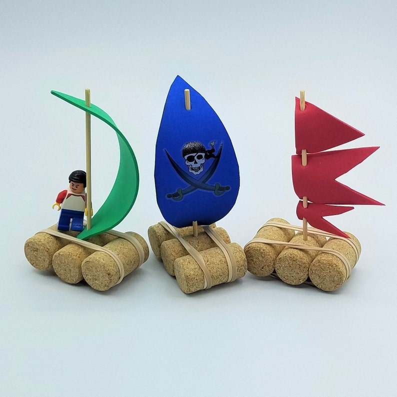 May include: Three miniature pirate rafts made from cork and decorated with colorful paper sails. One raft features a green sail, another a blue sail with a skull and crossbones, and the third a red sail. A small Lego figure stands on the raft with the green sail.
