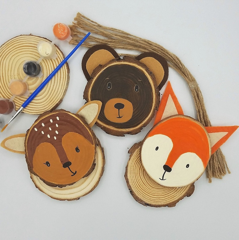 wood slice ornaments painted with the faces of a deer, bear, and fox, a paint strip,paint brush,and bundle of twine are placed around them