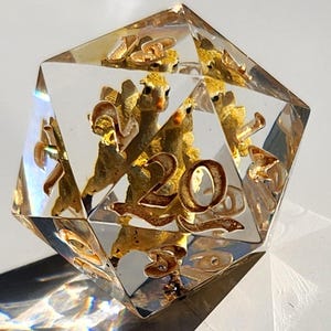 May include: A clear, multi-faceted D20 die with gold-colored llama figures and the number 20. The die is made of transparent resin, with the llama figures and number embedded inside. The die casts a shadow on a white surface.
