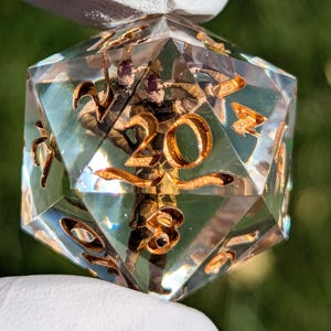 May include: A clear, multi-sided dice with gold numbers, 1 through 20, embedded inside. The dice is made of resin and has a clear, glassy appearance.
