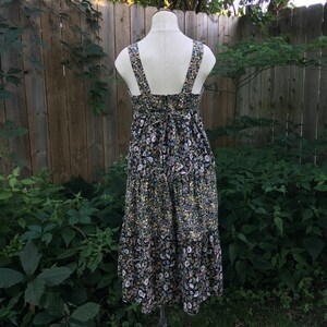 Puede incluir: Vestido midi con estampado floral sobre un maniqu&iacute;. El vestido presenta un fondo negro con un estampado de peque&ntilde;as flores blancas, azules y amarillas. Tiene un escote cuadrado, tirantes anchos y una falda escalonada. El vestido se exhibe al aire libre.