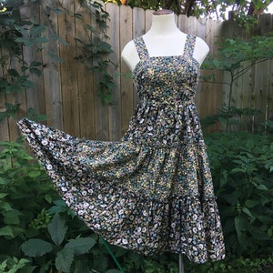 Puede incluir: Vestido con estampado floral sobre un maniqu&iacute;. El vestido presenta un fondo negro con un estampado de peque&ntilde;as flores blancas, amarillas y azules. Tiene un escote cuadrado, tirantes anchos y una falda escalonada, ideal para el verano.