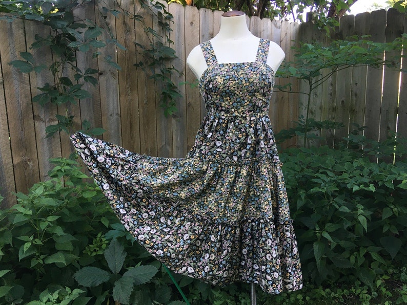 Puede incluir: Vestido con estampado floral sobre un maniqu&iacute;. El vestido presenta un fondo negro con un estampado de peque&ntilde;as flores blancas, amarillas y azules. Tiene un escote cuadrado, tirantes anchos y una falda escalonada, ideal para el verano.