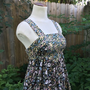 Puede incluir: Vestido de verano con estampado floral sobre un maniqu&iacute;. El vestido presenta un fondo negro con un estampado de flores blancas, azules y amarillas. Tiene un escote cuadrado, tirantes anchos y cintura fruncida. El vestido se exhibe al aire libre.